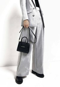 Czarna skórzana torba o zwartej formie z górnym uchwytem, wyposażona w odpinany pasek na ramię. Na tle jasnoszarego garnituru.