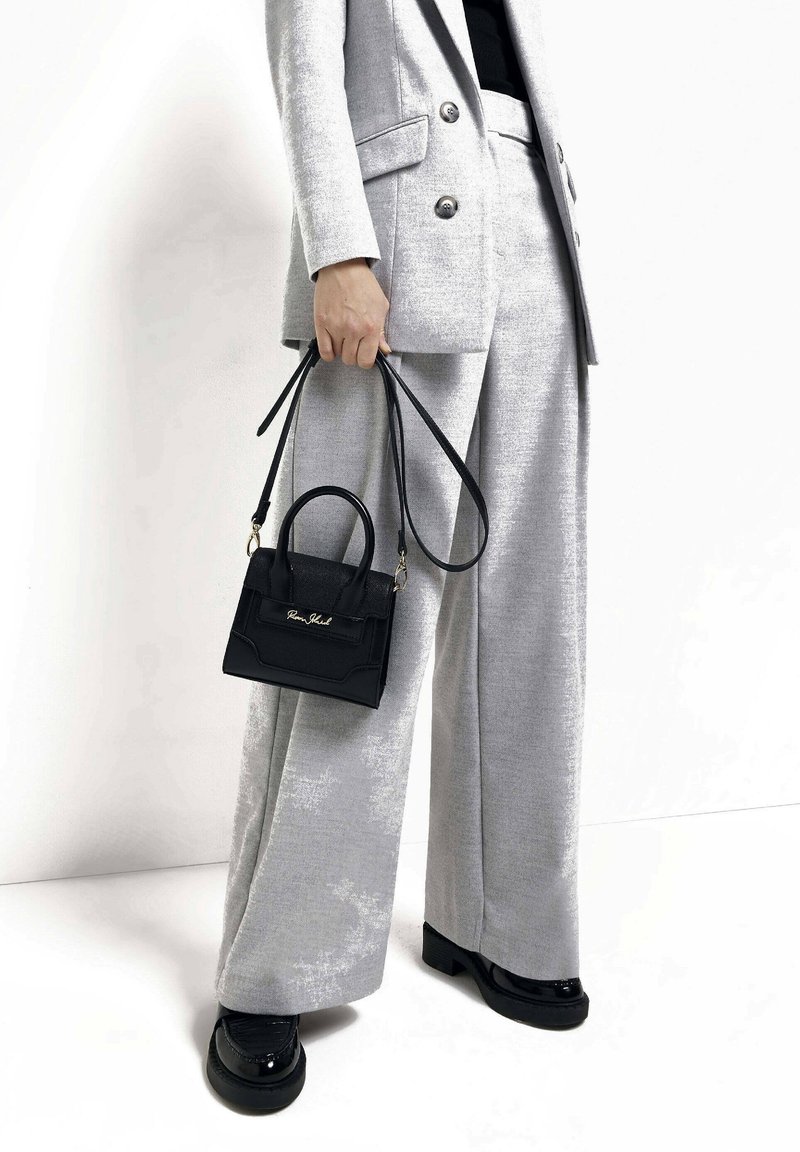 Czarna skórzana torba o zwartej formie z górnym uchwytem, wyposażona w odpinany pasek na ramię. Na tle jasnoszarego garnituru.