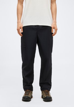 Mann trägt schwarze Cargohosen, weißes T-Shirt und braune Wanderschuhe, steht vor einem schlichten Hintergrund.