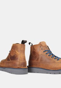 Scarpe alta in pelle marrone con finitura testurizzata, lacci blu, dettaglio esagonale e suole in gomma grigia, dotate di un pull tab sul tallone.