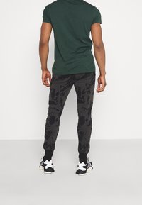 Donkergroene korte mouwen shirt gecombineerd met zwarte joggers met camo-patroon, voorzien van elastische mouwen en witte sneakeraccenten.