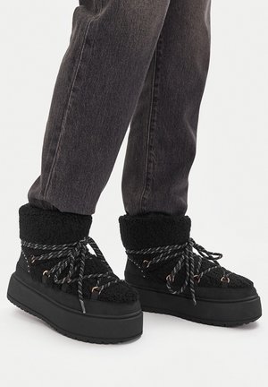 Czarne zimowe buty na platformie z grubymi dzianinowymi mankietami i odblaskowymi sznurówkami, noszone z ciemnoszarymi jeansami.