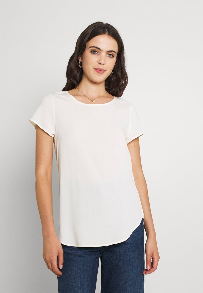 Hvit kortermet bluse med rund hals, myk tekstur, avrundet nederkant og en avslappet passform, kombinert med blå denimjeans.