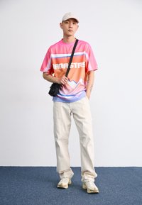 Colorful short-sleeve t-shirt with gradient pink and orange design, featuring bold white text. Paired with cream striped pants and beige sneakers.