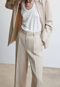 Béžový tailored blazer a kalhoty s širokými nohavicemi vyrobené z texturované tkaniny, kombinované s bílým tílkem. Oblečení má čisté linie a minimalistický design.