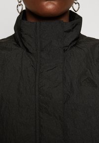 Svart Adidas-jacka med en krinklad textur, hög krage och subtil logotypdetalj. Har en elegant, minimalistisk design.