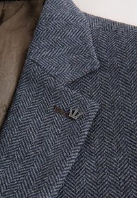 Dunkelblaues und graues Fischgrätenmuster-Sakko-Revers mit einer kleinen silbernen, kronenförmigen Anstecknadel nahe dem Knopfloch.