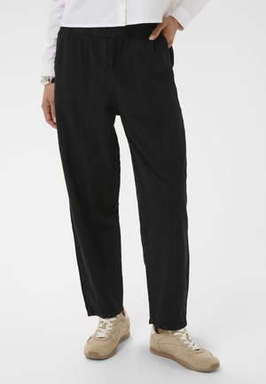 Person iført sorte bukser med løs pasform og lommer, beige sneakers og en hvid trøje med lange ærmer, stående mod en hvid baggrund.