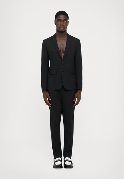 Zwart maatpak met een diepe V-hals blazer, rechte broek en glanzende zwarte instappers met gouden accenten. Eenvoudig, gestroomlijnd ontwerp.