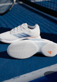 Λευκά αθλητικά παπούτσια με ανοιχτό μπλε ρίγες, υφασμένο καουτσούκ στις σόλες και πορτοκαλί λεπτομέρειες, τοποθετημένα σε μπλε επιφάνεια γηπέδου.