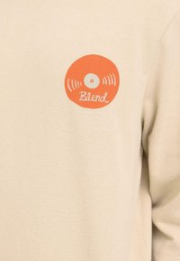 Chemise beige à manches longues en tissu doux, ornée d'un logo disque vinyle orange avec le mot "Blend" en écriture blanche cursive.