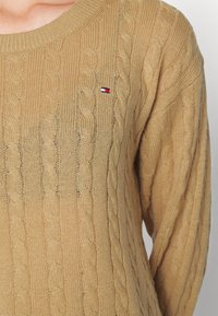 Beiger Zopfmuster-Pullover mit rundem Halsausschnitt, langen Ärmeln und einem kleinen gestickten Logo auf der Brust. Die strukturierte Stoffoberfläche verleiht dem Kleidungsstück Tiefe.