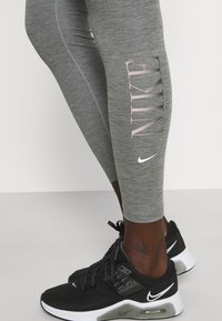 Šedé legíny s velkým růžovým logem "NIKE" na boku, doplněné černými sportovními botami se bílými akcenty a texturovaným materiálem.