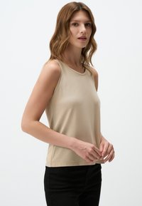 Top beige senza maniche realizzato in tessuto morbido con scollatura rotonda e silhouette aderente, abbinato a pantaloni neri. Design semplice, dettagli minimi.