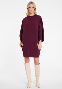 Lila Shiftkleid mit weiten Ärmeln, zwei vorderen Taschen, verziert mit goldenen Akzenten, getragen mit beigefarbenen knielangen Stiefeln, glatte Stoffstruktur.