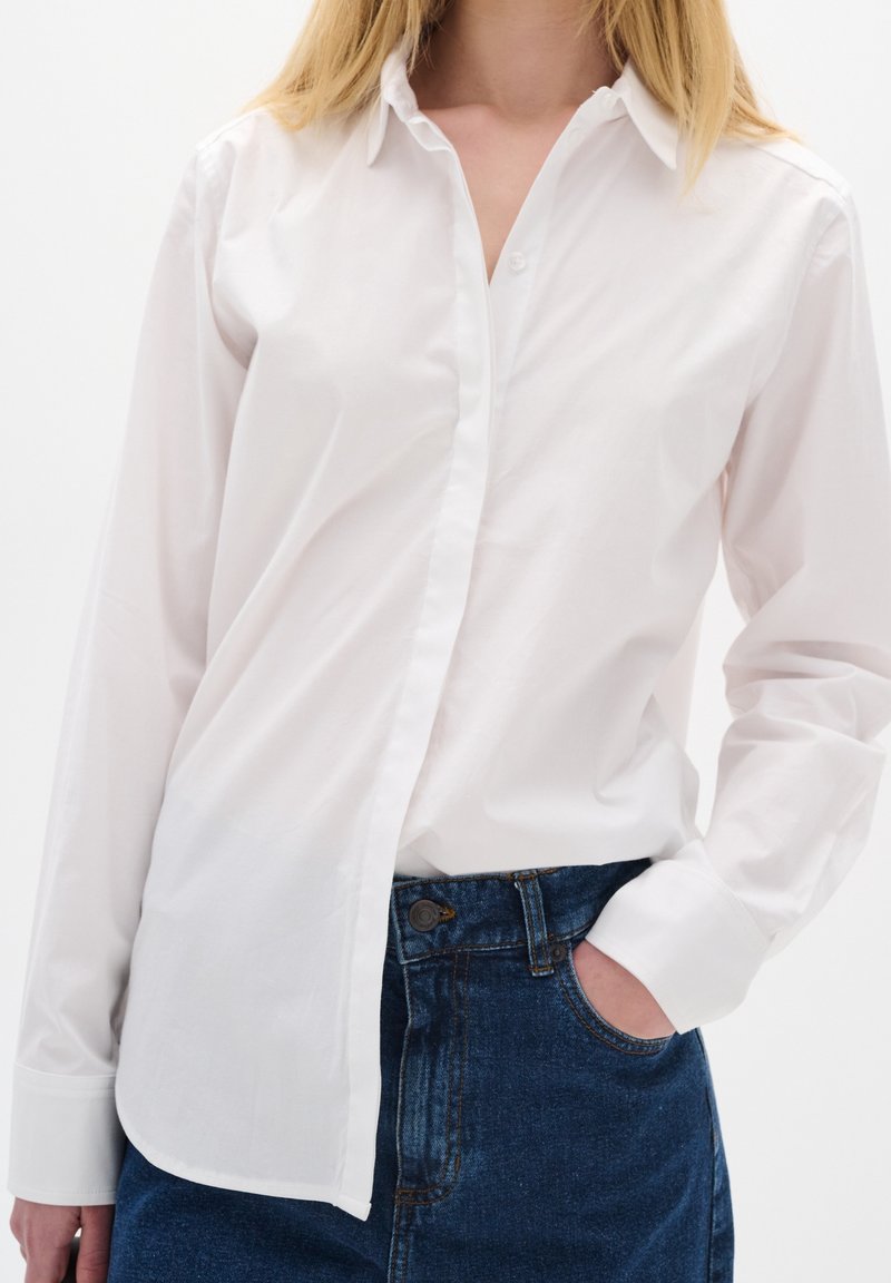 Camisa blanca de botones hecha de algodón con cuello clásico y mangas largas, combinada con jeans de mezclilla de talle alto. Textura suave, sin patrones.