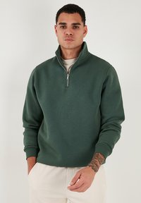 Pullover verde a mezza zip realizzato in tessuto morbido, con maniche lunghe, colletto a coste e design minimalista. Tessuto liscio e vestibilità comoda.