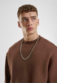 Sudadera marrón con un escote redondeado, que presenta un collar de cadena plateada brillante. La tela parece suave y ligeramente texturizada.