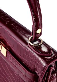 Mörk burgundy handväska i texturerat konstläder med krokodilsmönster. Guldhardwaredetaljer och ett handtag på toppen med sydda detaljer.