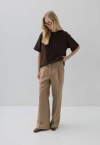 Brązowa oversize t-shirt zestawiona z beżowymi spodniami o wysokim stanie. Modelka nosi szare okulary przeciwsłoneczne i brązowe obuwie na jednolitym tle.