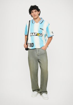 Polo a righe blu chiaro e bianche con logo nero "NAPOLI" davanti e numero 10, abbinato a jeans grigi sbiaditi e larghi e scarpe da ginnastica bianche.