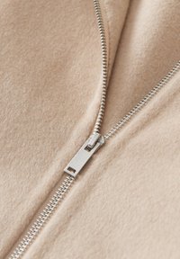 Beiger Fleece-Stoff mit einem auffälligen silbernen Reißverschluss. Nahaufnahme hebt die Textur und das glatte, weiche Material hervor. Der Reißverschluss ist gut sichtbar.