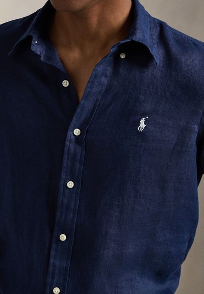 Hombre que lleva una camisa de lino azul marino abotonada con botones blancos y un logo bordado blanco de un jugador de polo en el pecho.