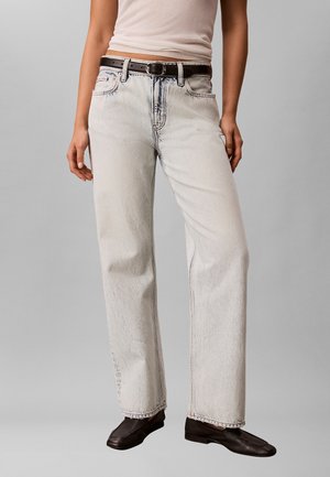 Person iført lysvaskede straight-leg jeans med sort bælte, beige ærmeløs top og sorte flade sko, stående mod en grå baggrund.