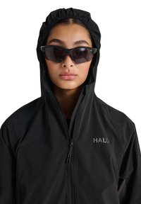 Person trägt eine schwarze Kapuzenjacke mit „HALO“-Logo und schwarze, eng anliegende Sonnenbrille vor weißem Hintergrund.