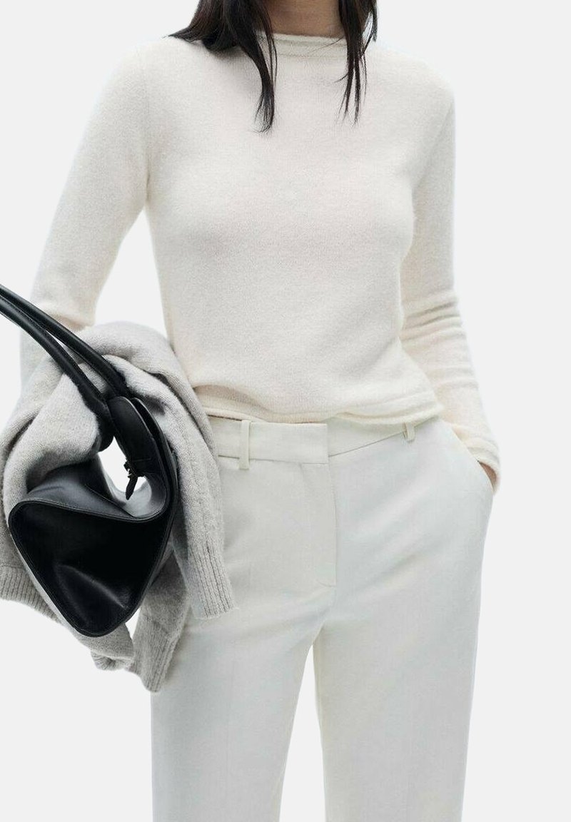 Femme portant un pull blanc à manches longues et un pantalon blanc, tenant un pull gris et un sac à main noir, une main dans la poche.
