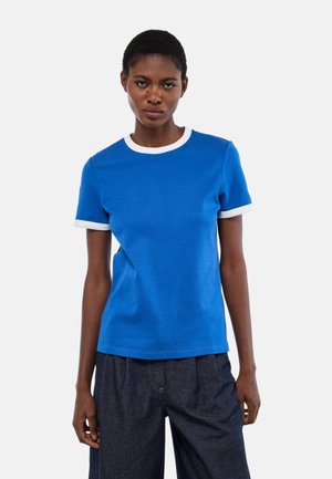 Femme portant un t-shirt bleu avec col et bords de manches blancs, associé à un pantalon ample foncé, se tenant devant un fond uni.
