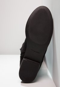 Semelle de chaussure noire avec une texture antidérapante et un petit talon, reposant verticalement contre une surface blanche.