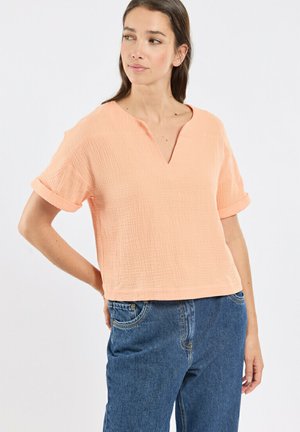 Femme portant un haut texturé à manches courtes de couleur orange clair avec un décolleté en encoche et un jean bleu taille haute, regardant sur le côté.