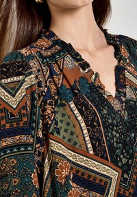 Blusa a fantasia con una combinazione di verde scuro, marrone e blu. Collo arricciato, patta con bottoni e motivi floreali geometrici in tutto il capo.