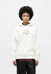 Krém színű kapucnis pulóver virágmintával és "Hugo" felirattal. Elöl zsebbel, zsinóros kapucnival és kényelmes szabással. Bordó felső fölött viselve.