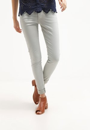 Vrouw die lichtgrijze skinny jeans draagt, opgerold bij de enkels, bruine open teen enkellaarsjes met hak, en een marineblauwe scalloped kanten top.