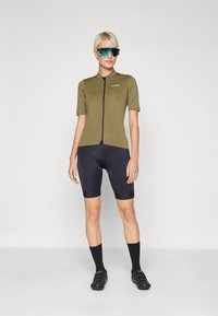 Jersey de ciclismo verde oliva de manga corta con cremallera frontal, combinado con pantalones cortos acolchados negros y calcetines negros, usado con gafas de sol azules.