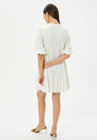 Robe blanche à manches courtes avec un tissu texturé, un ourlet froncé et une coupe décontractée. Portée avec des sandales à talons dorés, vue de dos.