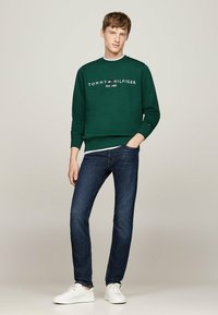 Sweatshirt en coton vert avec le logo blanc 'TOMMY HILFIGER', associé à un jean bleu marine et des baskets blanches, se tenant devant un fond neutre.