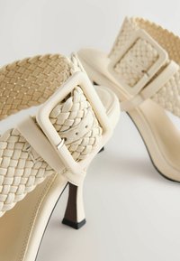 Pletené kožené sandály v off-white barvě, se čtvercovou sponou a jehlovým podpatkem s černým špičkou. Texturované popruhy přidávají vizuální zajímavost.