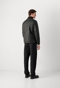 Veste matelassée vert olive avec un col marron foncé, accents de boutons à l'ourlet, associée à un pantalon large gris foncé et des chaussures noires.