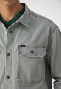 Camisa gris de botones con un bolsillo en el pecho, botones de cobre y un cuello de canalé verde. El material tiene una textura sutil. Etiqueta de la marca visible.