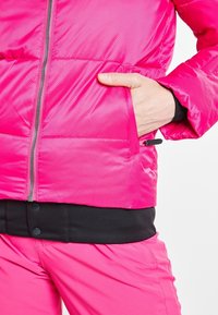 Veste matelassée rose vif avec une finition texturée, dotée d'une fermeture éclair frontale et d'une poche latérale. Détail de ceinture élastique noire en bas.