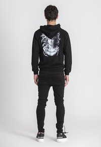 Crna dukserica s grafičkim otiskom lica okruženog bujnom kosom na leđima, u kombinaciji s crnim trapericama i bijelim tenisicama.