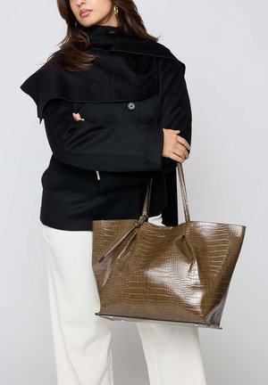 Vrouw met een zwarte jas en een witte broek die een grote bruine krokodillenleer tote bag met dubbele handvatten vasthoudt tegen een grijze achtergrond.