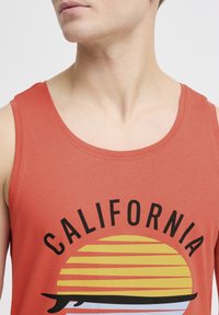 Tricou roșu fără mâneci cu un grafic care prezintă cuvântul "CALIFORNIA" și un design cu apus de soare, cu dungi galbene și portocalii și o siluetă neagră.