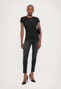 Zwarte getailleerde t-shirt met korte mouwen, voorzien van een gouden logo op de borst. Gecombineerd met donkergrijze skinny jeans en zwarte hoge hakken.