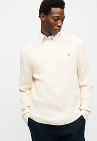 Mężczyzna ubrany w kremowy teksturowany sweter na białej koszuli z kołnierzykiem, z rękami w kieszeniach ciemnych spodni, patrzący w dół w lewo.