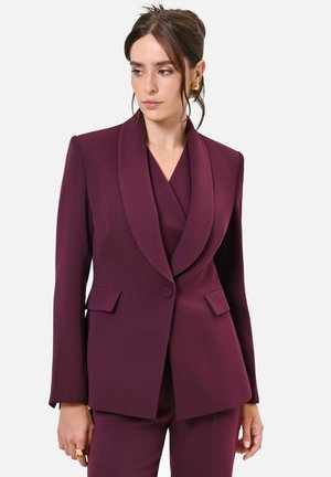 Donna che indossa un completo su misura colore viola scuro con blazer con collo a scialle e pantaloni coordinati, che guarda leggermente verso sinistra su uno sfondo semplice.