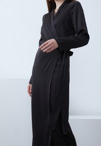 Robe noire en satin lisse avec des manches longues, un décolleté en V profond et un lien à la taille. Présente une silhouette longue jusqu'au sol.